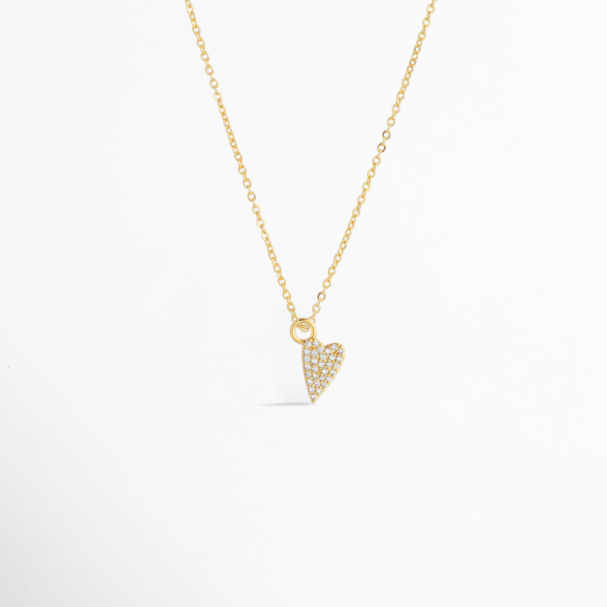 Charm Corazón Zirconias montado como dije central en una fina cadena dorada sobre fondo blanco, Vista de collar con dije pequeño de corazón estilizado enchapado en oro con circonias brillantes de 1.2 cm, Perspectiva de gargantilla delicada con dije colgante en forma de corazón dorado con pedrería, Muestra de escala del charm de corazón incrustado con circonias en una cadena delgada enchapada en oro.
