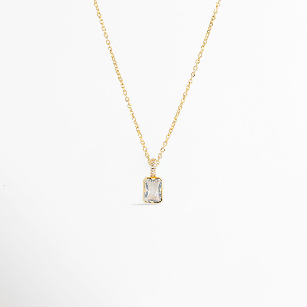 Charm Double Shimmer suspendido de una fina cadena dorada mostrando la proporción del dije rectangular en un collar sobre fondo blanco, Vista general de collar fino dorado enriquecido con el dije rectangular de circonia transparente Double Shimmer, Plano medio que resalta la caída elegante y la reasa brillante del charm de 1.1 cm en una cadena dorada, Muestra del brillo y la claridad del corte esmeralda sobre el escote en una composición de cadena fina.