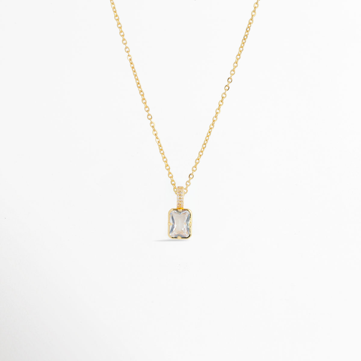 Charm Double Shimmer suspendido de una fina cadena dorada mostrando la proporción del dije rectangular en un collar sobre fondo blanco, Vista general de collar fino dorado enriquecido con el dije rectangular de circonia transparente Double Shimmer, Plano medio que resalta la caída elegante y la reasa brillante del charm de 1.1 cm en una cadena dorada, Muestra del brillo y la claridad del corte esmeralda sobre el escote en una composición de cadena fina.