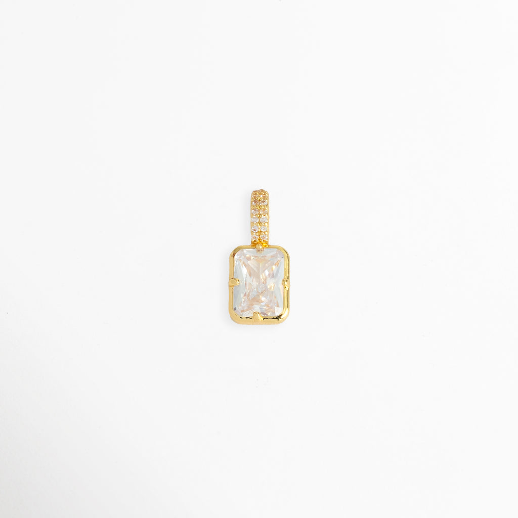 Charm Double Shimmer de cobre con acabado galvanizado dorado de 1.1 cm de largo y corte esmeralda en circonia transparente con reasa engastada, vista frontal sobre fondo blanco, Vista de frente de dije rectangular dorado con gran circonia cristalina central de corte radiante y reasa con pavé de pedrería, Plano general de charm elegante con gema transparente de corte esmeralda de 0.7 cm de ancho y bisel dorado de alto brillo, Charm Double Shimmer destacando el doble 