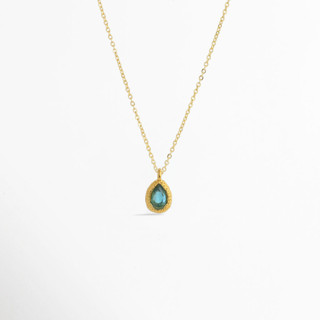 Charm Gota Azul Claro luciendo como colgante central en una fina cadena dorada, vista frontal en plano general, Collar dorado con dije en forma de gota azul claro de diseño sereno y elegante, Vista de colgante en tono azul mar sobre cadena fina enchapada en oro sobre fondo blanco, Ejemplo de estilismo del Charm Gota Azul Claro integrado como punto de color fresco en un collar dorado.