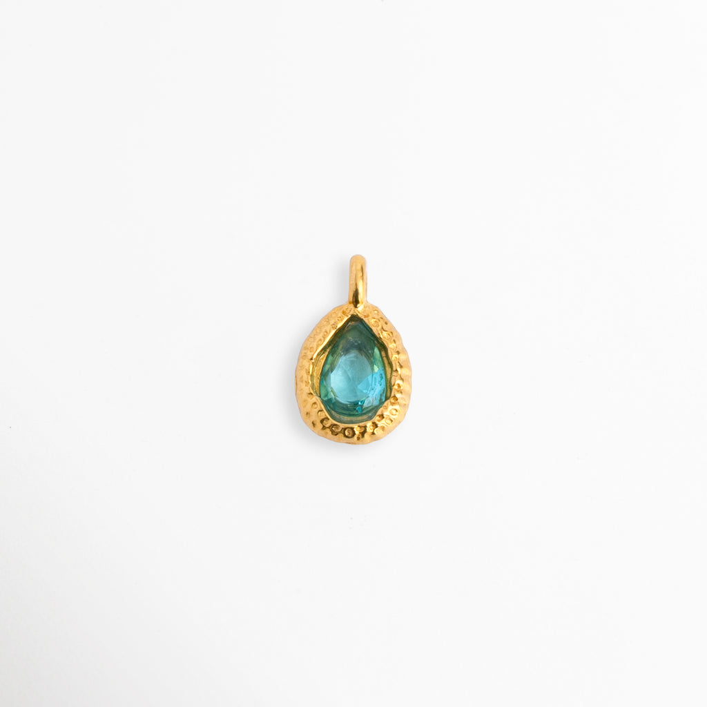 Charm Gota Azul Claro con circonia en tono azul cielo de corte lágrima y marco orgánico texturizado en tono dorado, vista frontal sobre fondo blanco,  Dije de 1.2 x 0.7 cm en forma de gota con gema azul claro engastada en acero inoxidable con baño de oro, Vista de frente de dije charm de acero dorado con circonia azul celeste de corte pera y borde artesanal, Charm de acero inoxidable enchapado en oro con piedra de circonia azul acuamarina sobre fondo claro.