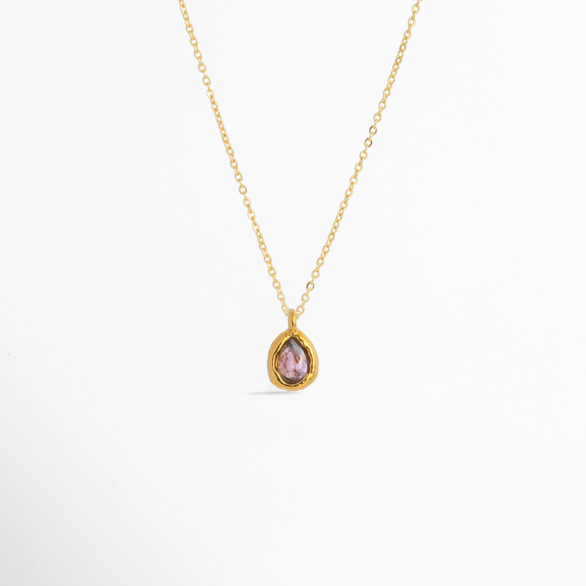Charm Gota Rosa luciendo como colgante central en una fina cadena dorada, vista frontal en plano general, Collar dorado con dije en forma de gota rosa de diseño delicado y femenino, Vista de colgante en tono rosa suave sobre cadena fina enchapada en oro sobre fondo blanco, Ejemplo de estilismo del Charm Gota Rosa integrado como punto de color en un collar dorado.