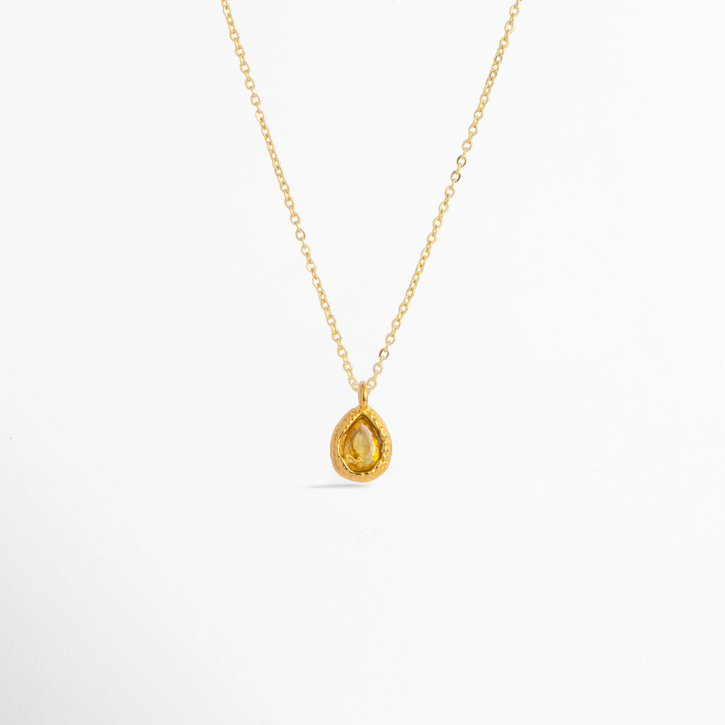 Charm Gota Amarillo luciendo como colgante central en una fina cadena dorada, vista frontal en plano general, Collar dorado con dije en forma de gota amarilla de diseño cálido y resplandeciente, Vista de colgante en tono amarillo sol sobre cadena fina enchapada en oro sobre fondo blanco, Ejemplo de estilismo del Charm Gota Amarillo integrado como punto de luz solar en un collar dorado.