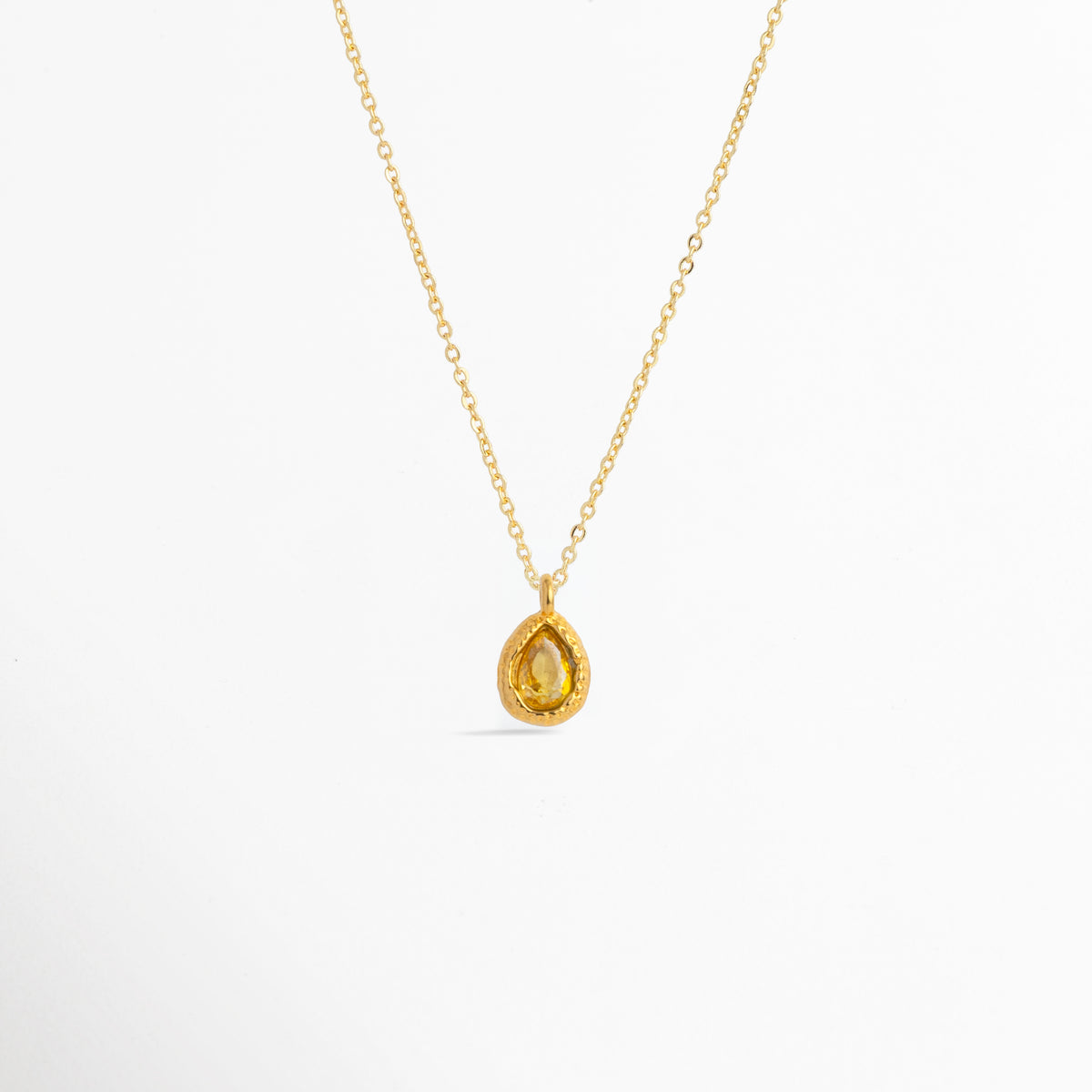 Charm Gota Amarillo luciendo como colgante central en una fina cadena dorada, vista frontal en plano general, Collar dorado con dije en forma de gota amarilla de diseño cálido y resplandeciente, Vista de colgante en tono amarillo sol sobre cadena fina enchapada en oro sobre fondo blanco, Ejemplo de estilismo del Charm Gota Amarillo integrado como punto de luz solar en un collar dorado.