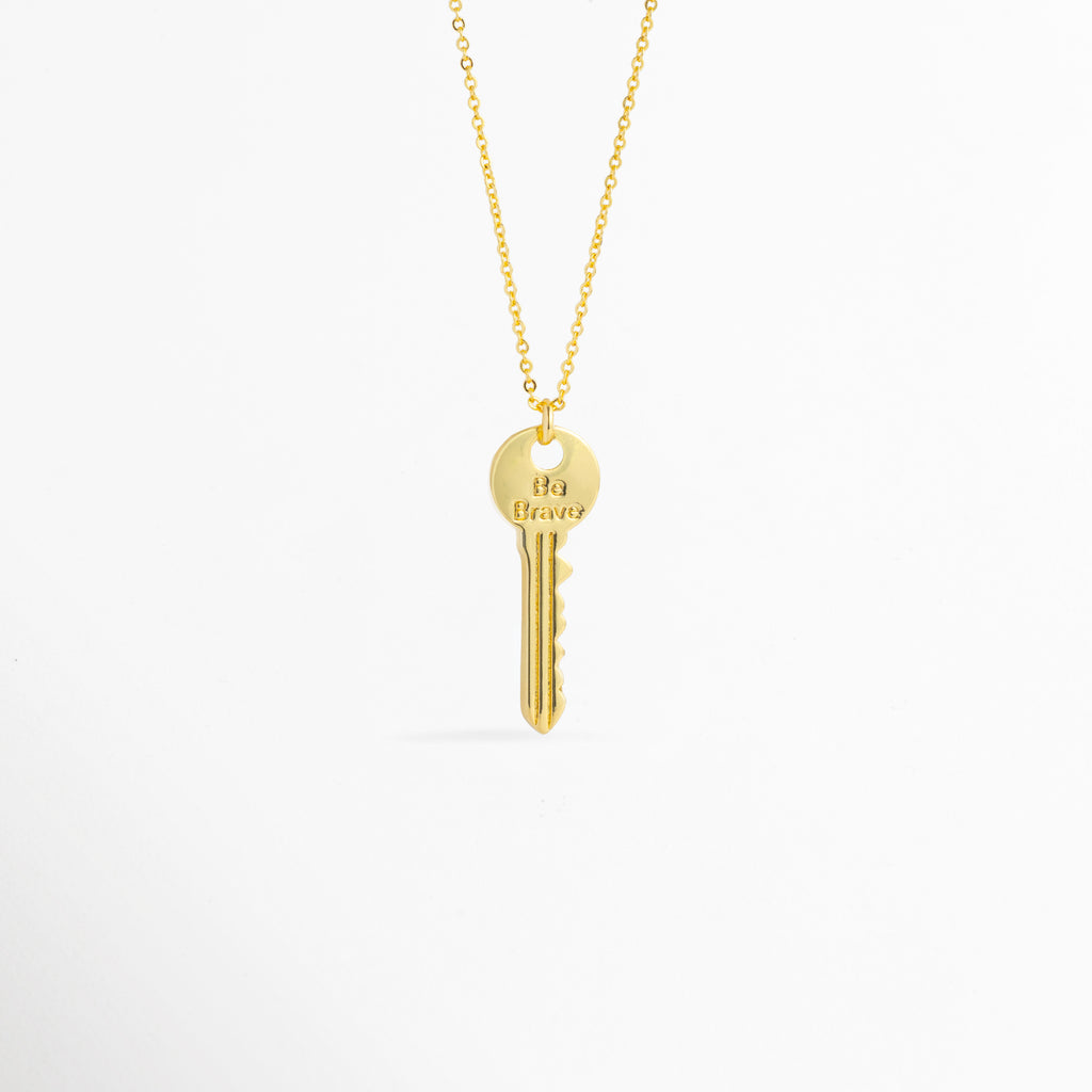 Charm Llave Be Brave de cobre enchapado en oro con grabado frontal de la frase Be Brave en la cabeza circular, vista de frente sobre fondo blanco, Vista frontal de dije en forma de llave clásica dorada de 3.5 cm de largo colgado de una fina cadena metálica, Plano general de charm estilo llave vintage enchapado en oro con mensaje inspirador grabado en la parte superior, Charm Llave Be Brave destacando los relieves de la dentadura de la llave