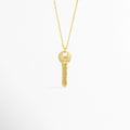 Charm Llave Be Brave de cobre enchapado en oro con grabado frontal de la frase Be Brave en la cabeza circular, vista de frente sobre fondo blanco, Vista frontal de dije en forma de llave clásica dorada de 3.5 cm de largo colgado de una fina cadena metálica, Plano general de charm estilo llave vintage enchapado en oro con mensaje inspirador grabado en la parte superior, Charm Llave Be Brave destacando los relieves de la dentadura de la llave