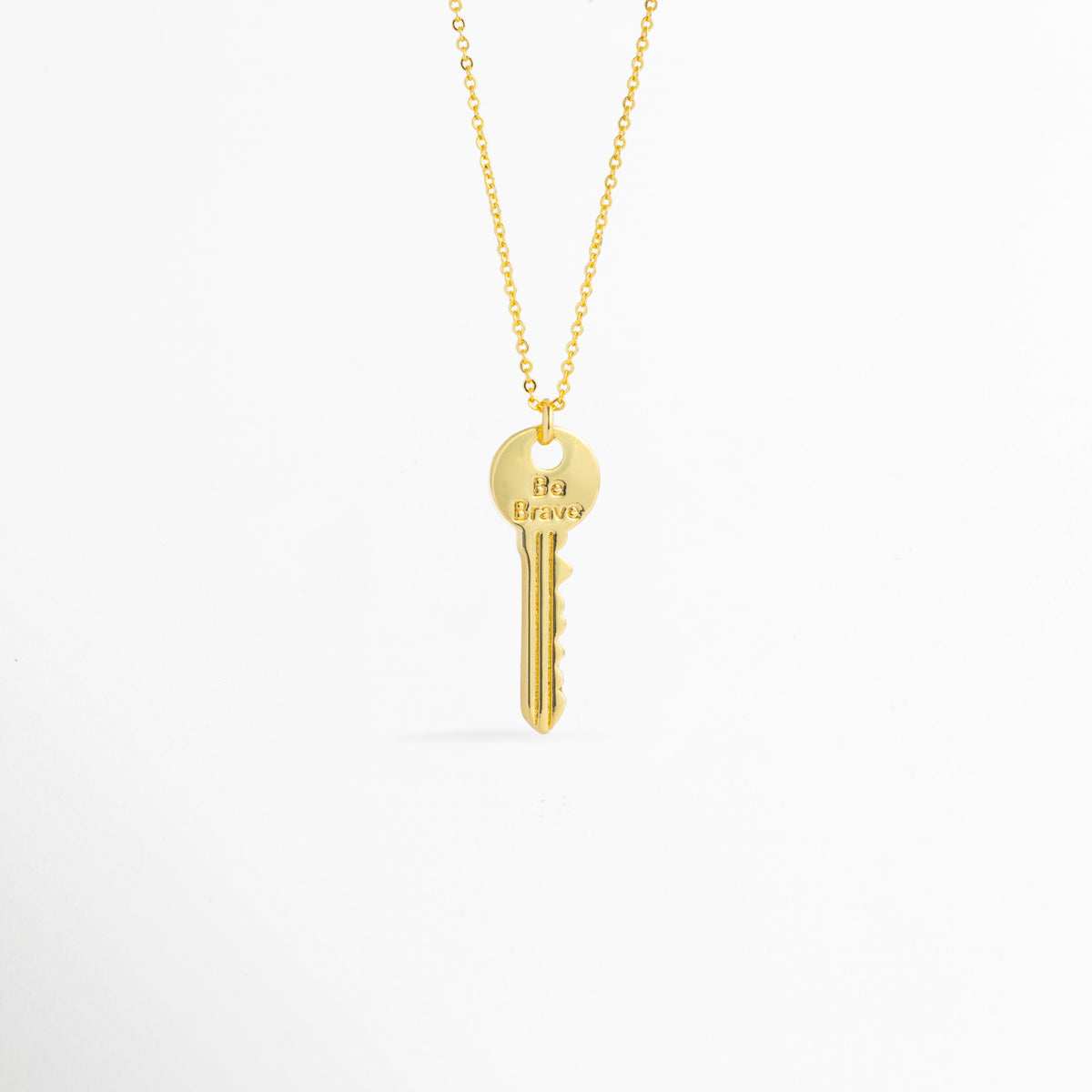 Charm Llave Be Brave de cobre enchapado en oro con grabado frontal de la frase Be Brave en la cabeza circular, vista de frente sobre fondo blanco, Vista frontal de dije en forma de llave clásica dorada de 3.5 cm de largo colgado de una fina cadena metálica, Plano general de charm estilo llave vintage enchapado en oro con mensaje inspirador grabado en la parte superior, Charm Llave Be Brave destacando los relieves de la dentadura de la llave