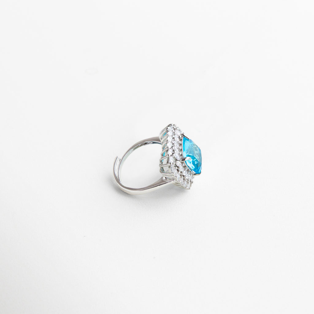 Anillo Lanikai Aqua visto de perfil, con borde plateado y piedra azul elevada que capta la luz con cada ángulo. Vista lateral del anillo con centro azul, mostrando su estructura delicada y brillo sofisticado. Perfil del anillo Lanikai Aqua, destacando el volumen de la zirconia azul y el contraste con el metal plateado. Detalle lateral del anillo con piedra azul, ideal para looks glamorosos y ocasiones especiales. Anillo Lanikai Aqua visto desde el lado, con diseño moderno y piedra central qu