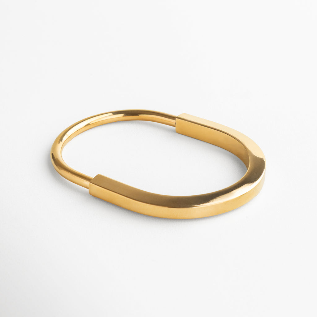 Pulsera dorada de acero inoxidable, Diseño estructurado y envolvente, Líneas limpias y definidas, Acabado brillante en baño de chapa de oro 18K, Forma ovalada para ajuste cómodo, Dimensiones: 7.3 cm largo, 5.8 cm ancho, Estilo moderno y sofisticado, ideal para outfits elegantes