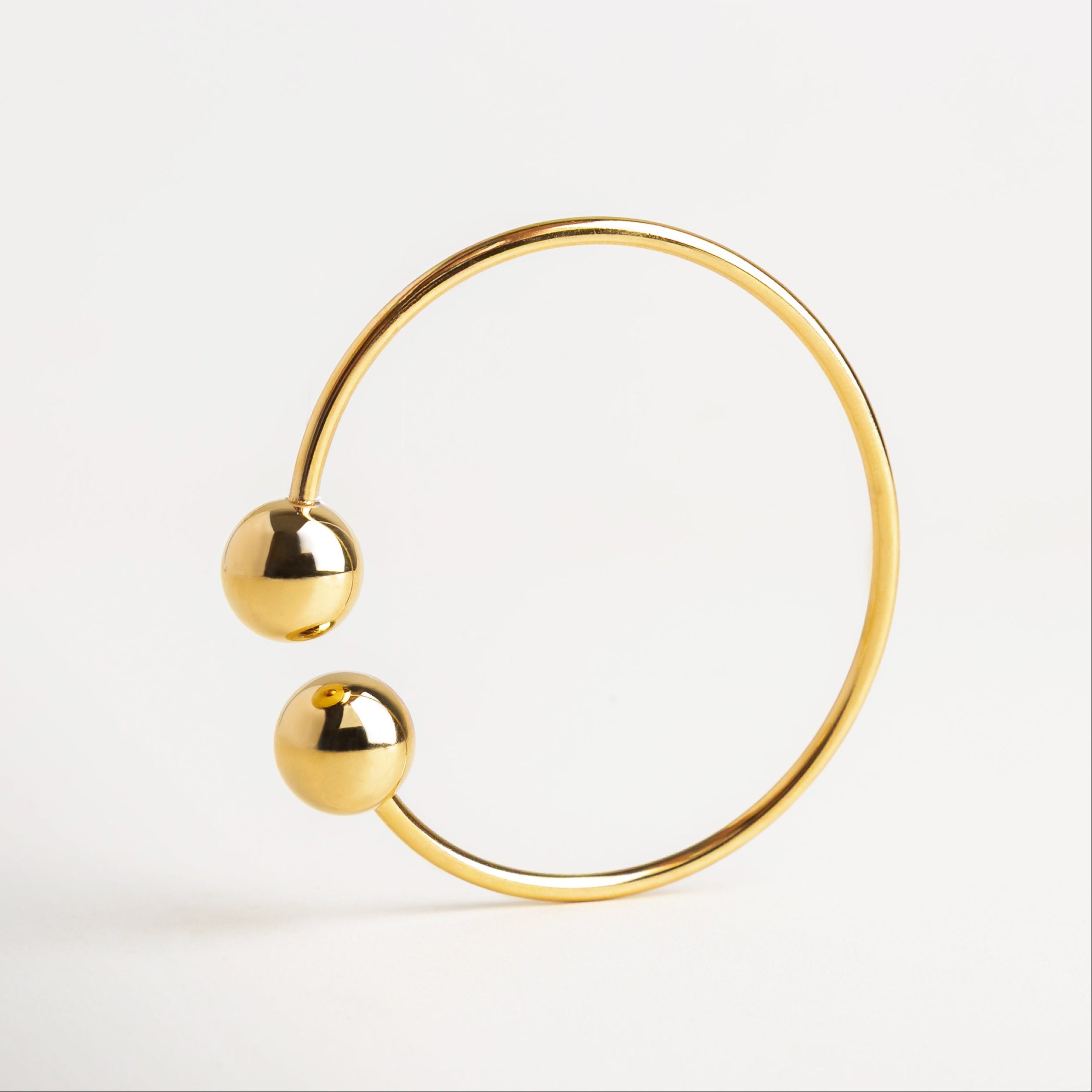 Pulsera Bensi dorada vista en ángulo moderno, Pulsera con diseño curvo y esferas terminales, Línea abierta de pulsera con estructura en dorado, Pulsera estructurada con esferas metálicas en dorado