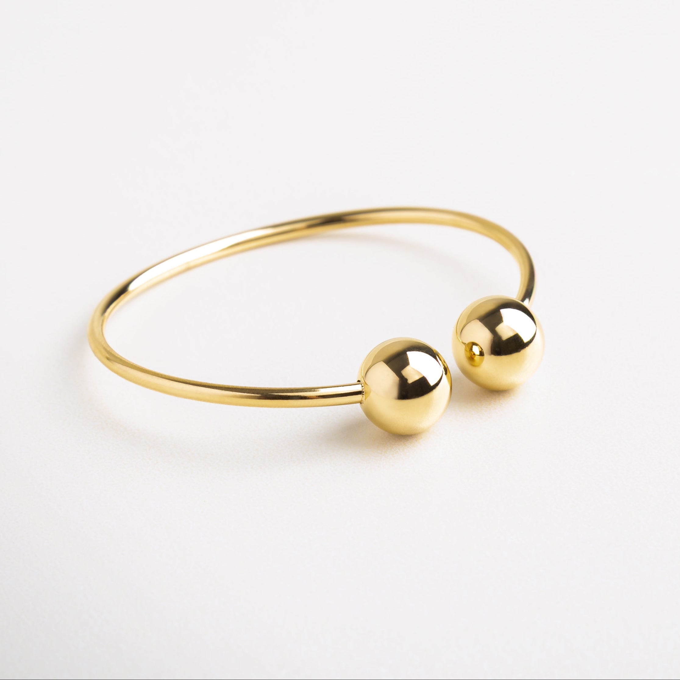 Detalle de esferas doradas en pulsera abierta, Acabado brillante en pulsera dorada minimalista, Textura suave y esferas pulidas en extremos de pulsera, Cierre abierto con diseño limpio en pulsera dorada