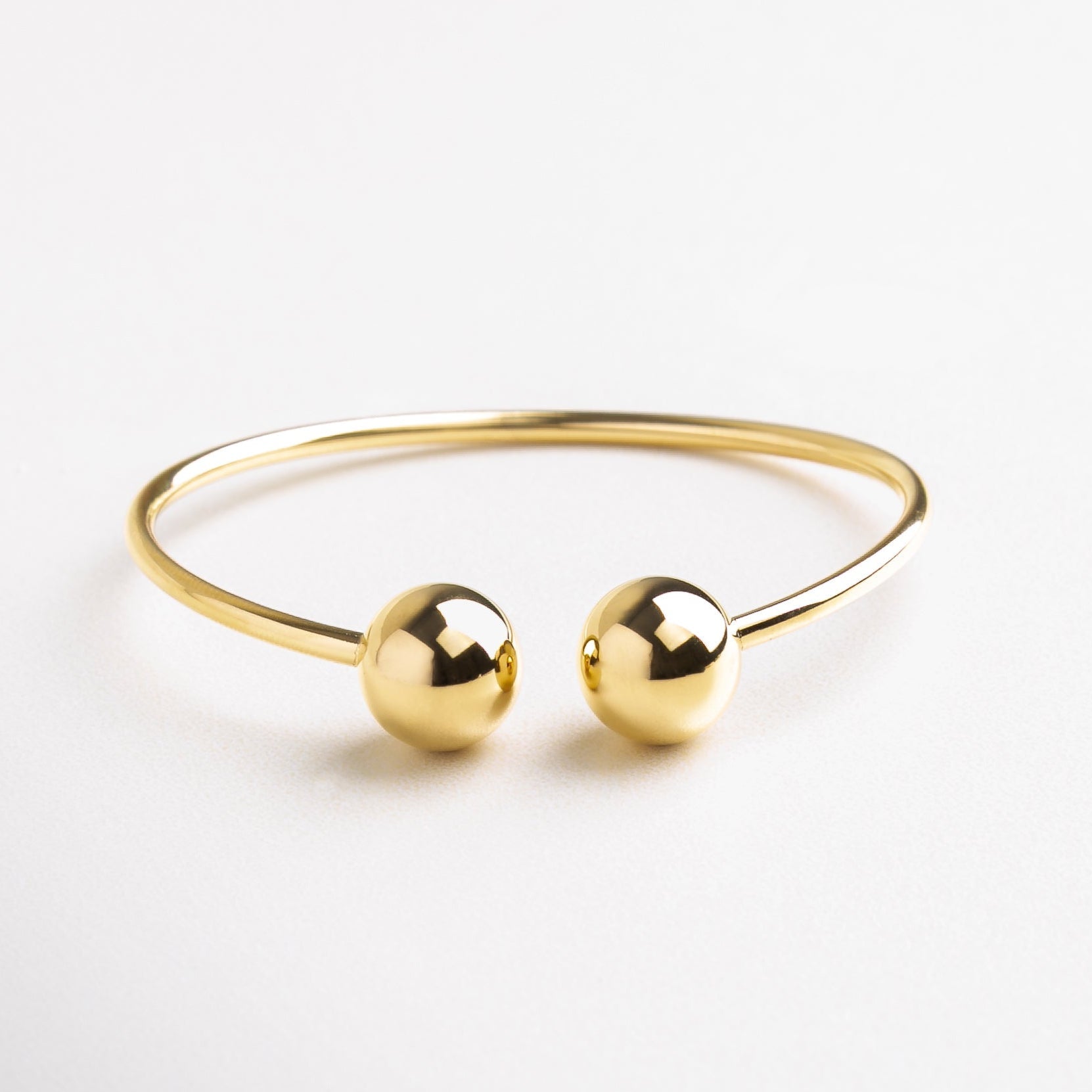 Pulsera dorada abierta con esferas en los extremos, Pulsera minimalista en dorado con diseño moderno, Pulsera de acero con acabado en chapa de oro 18K, Diseño abierto de pulsera dorada con detalles elegantes