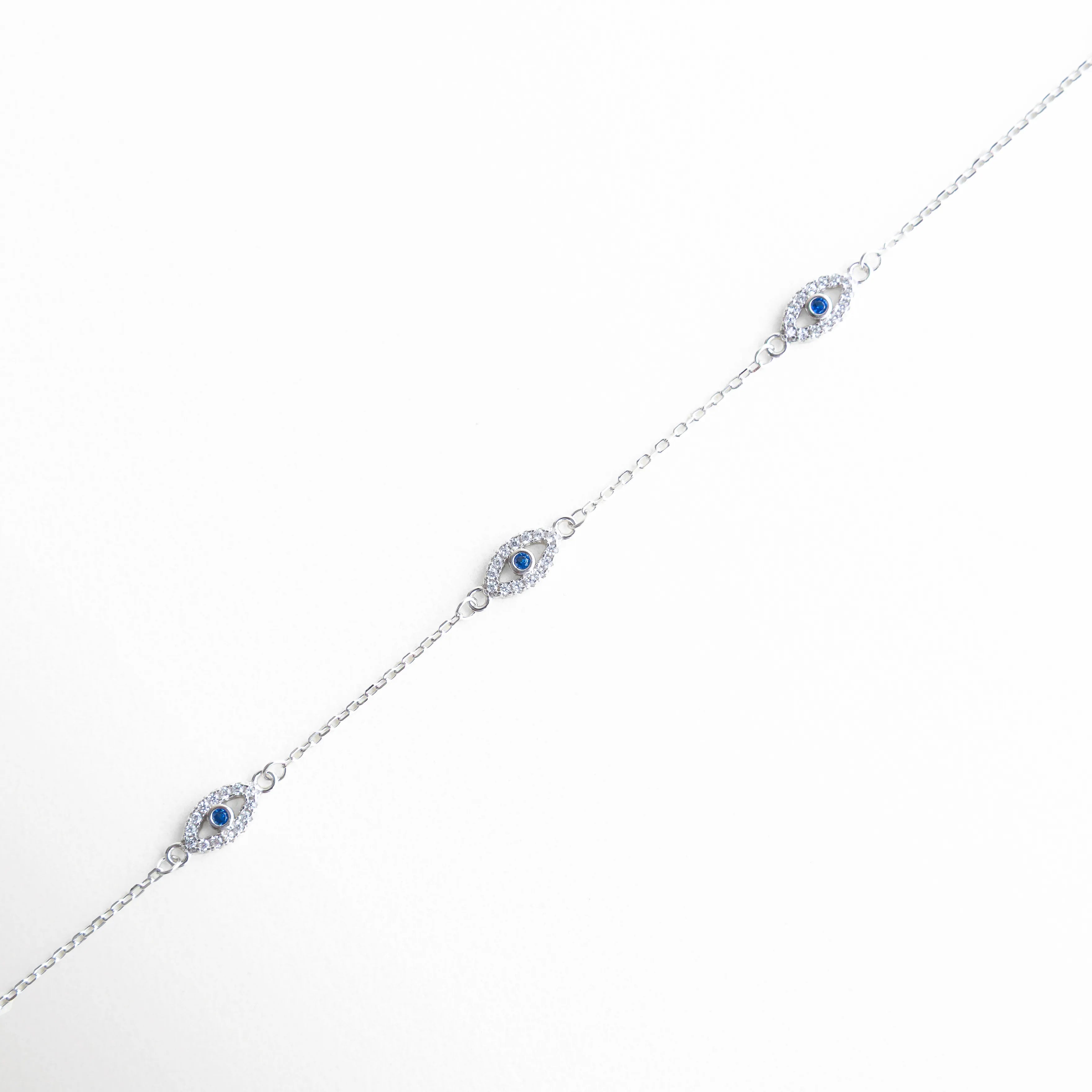 Pulsera plata 925 con tres ojos turcos y zirconias en acabado oro blanco 18K, Pulsera delicada con dijes de ojo turco azul y brillo sutil en fondo claro, Pulsera fina con tres amuletos ojo turco y zirconias alineadas en diagonal, Diseño elegante de pulsera plateada con ojos turcos y detalles brillantes, Pulsera minimalista con símbolos de protección y zirconias en tono plata