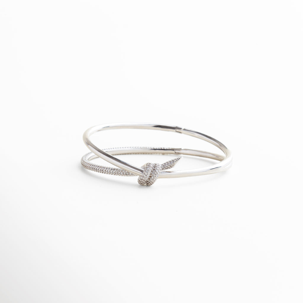 pulsera rígida plateada con diseño de nudo, brazalete plateado con zirconias incrustadas, diseño elegante con estructura abierta y minimalista, pulsera tipo statement con lazo brillante al centro, joya contemporánea en tono plateado con brillo sutil