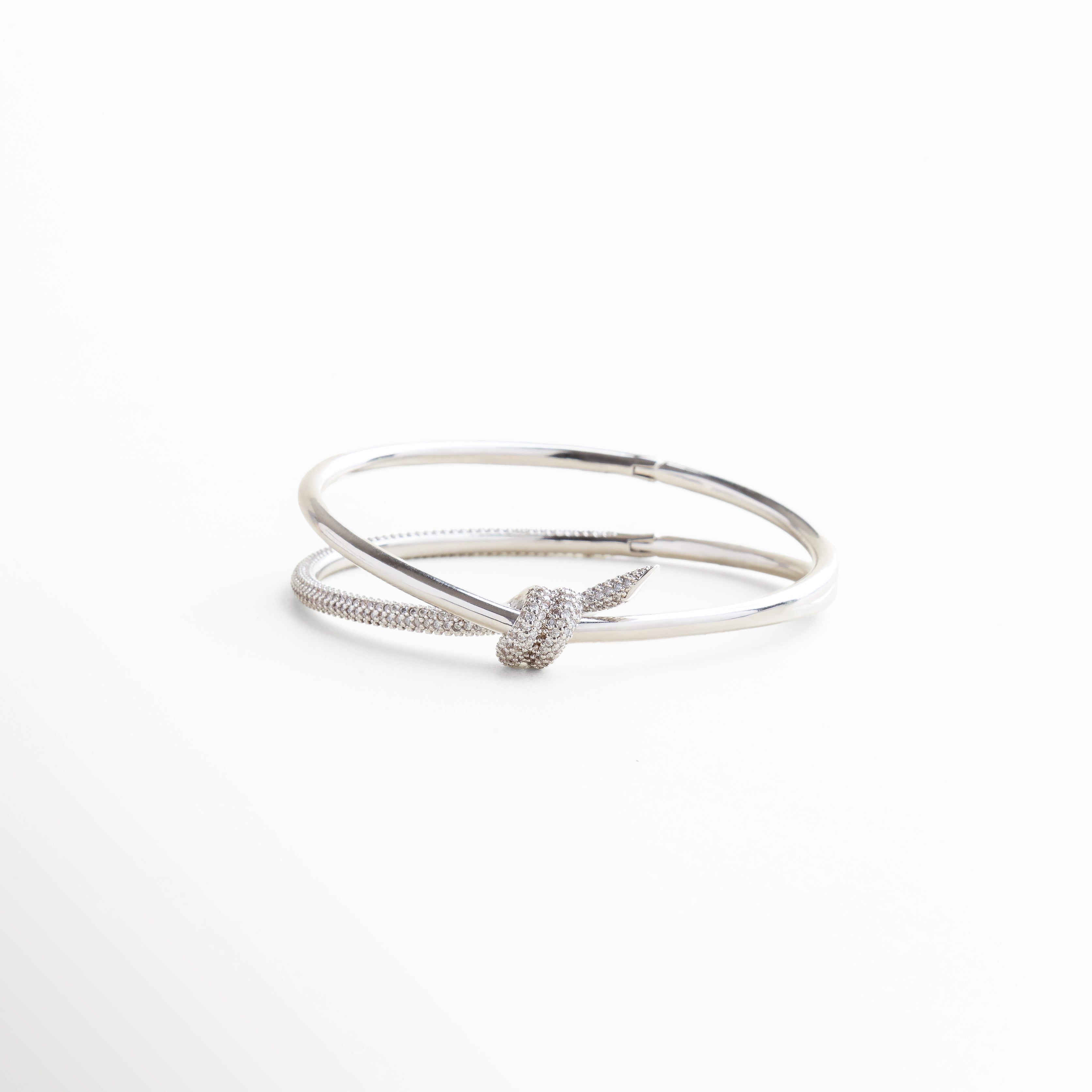 pulsera rígida plateada con diseño de nudo, brazalete plateado con zirconias incrustadas, diseño elegante con estructura abierta y minimalista, pulsera tipo statement con lazo brillante al centro, joya contemporánea en tono plateado con brillo sutil