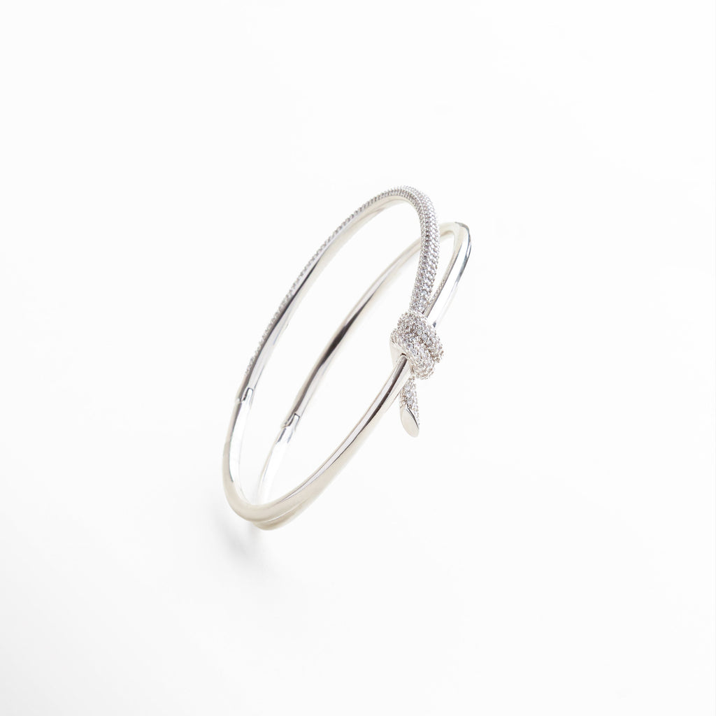 nudo central con incrustaciones de zirconias, acabado pulido con reflejo metálico brillante engarzado delicado de zirconias sobre el nudo textura lisa y líneas limpias del brazalete, detalle del cierre abierto y forma ovalada 