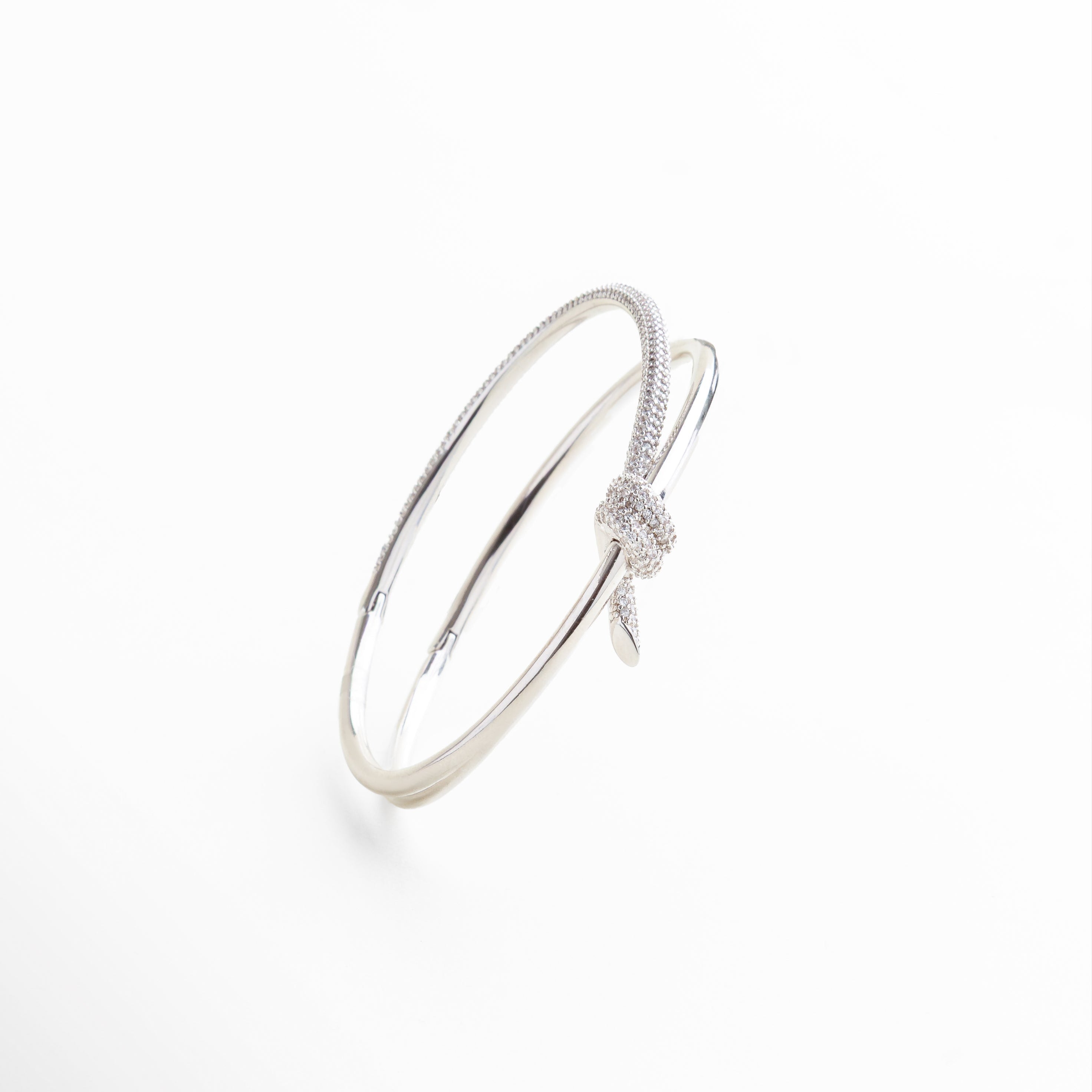 nudo central con incrustaciones de zirconias, acabado pulido con reflejo metálico brillante engarzado delicado de zirconias sobre el nudo textura lisa y líneas limpias del brazalete, detalle del cierre abierto y forma ovalada 