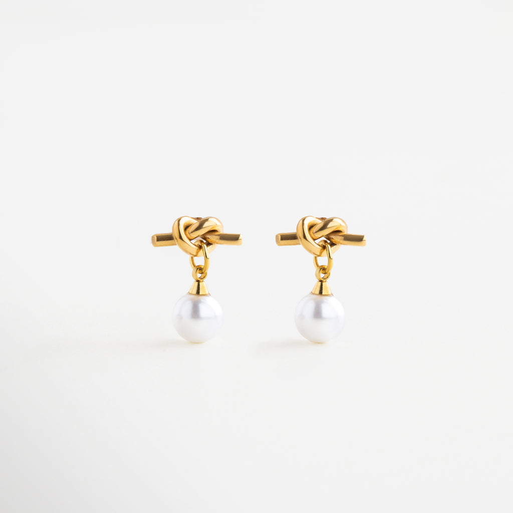 Aretes dorados con perla sintética y diseño de nudo, Aretes Knotté con baño de oro 18K y perla blanca, Aretes tipo nudo con perla, símbolo de unión y elegancia, Aretes modernos con perla sintética en acabado dorado