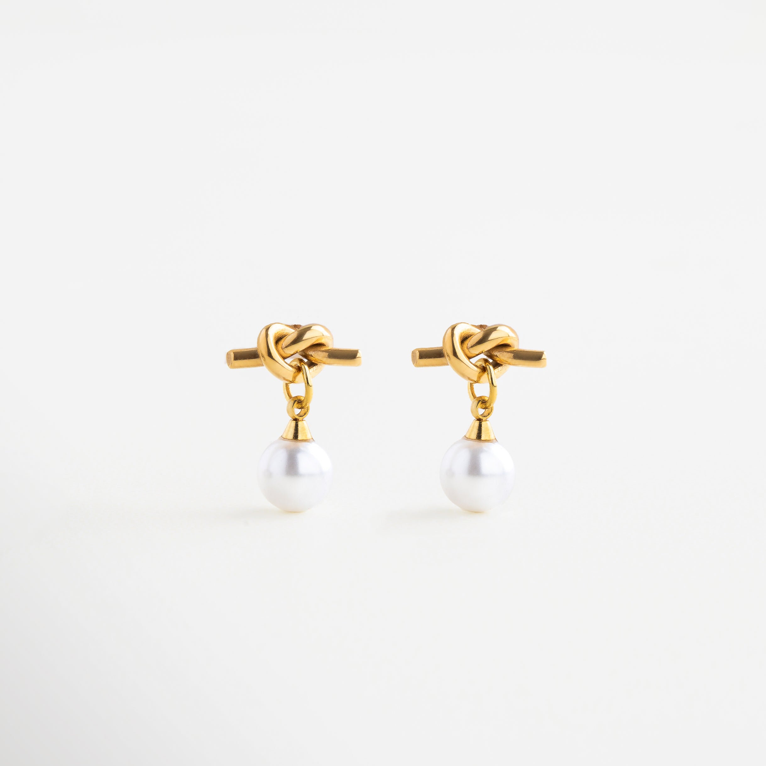 Aretes dorados con perla sintética y diseño de nudo, Aretes Knotté con baño de oro 18K y perla blanca, Aretes tipo nudo con perla, símbolo de unión y elegancia, Aretes modernos con perla sintética en acabado dorado