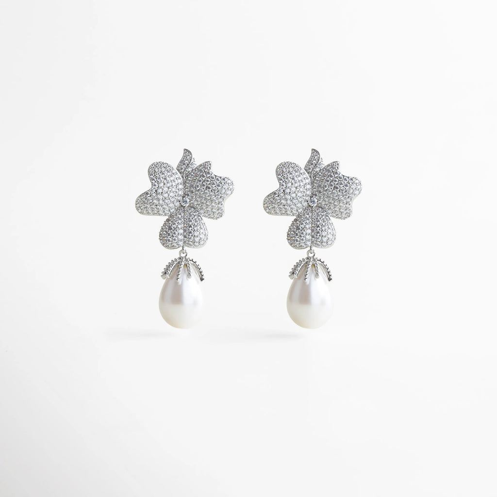 Par de aretes plateados con forma de flor y perla blanca colgante, diseño glamuroso y femenino que combina brillo y elegancia atemporal.