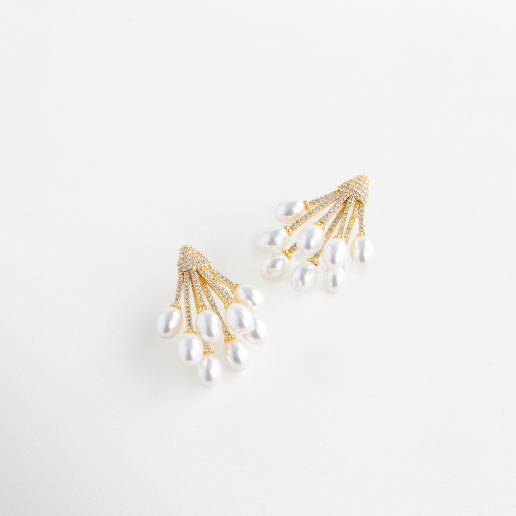 Vista lateral de aretes dorados con perla y zirconias brillantes, Aretes dorados con diseño curvo y perla sintética vistos en ángulo, Joyería dorada elegante con perla en movimiento y toques brillantes, Diseño lateral de aretes dorados con perla suspendida
