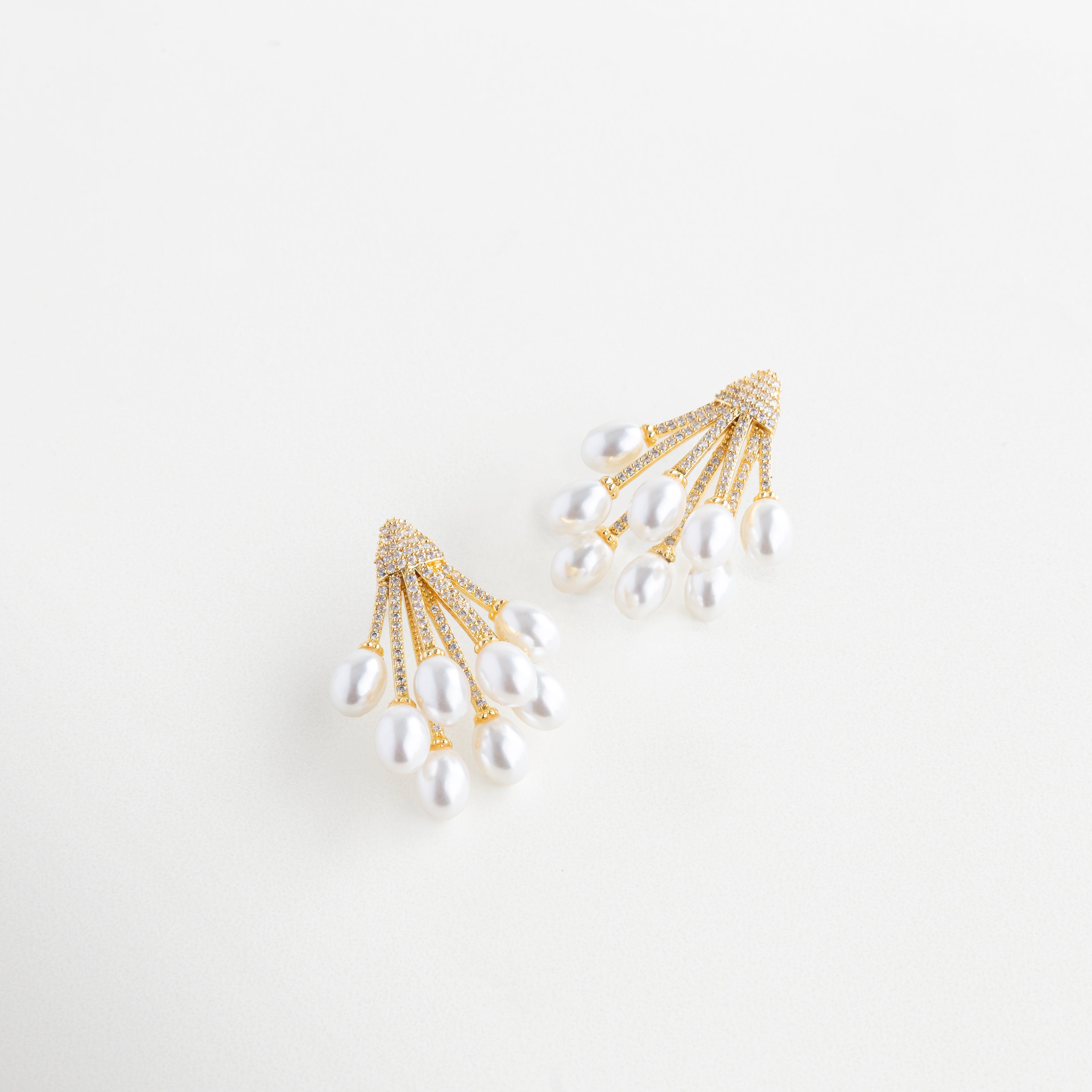 Vista lateral de aretes dorados con perla y zirconias brillantes, Aretes dorados con diseño curvo y perla sintética vistos en ángulo, Joyería dorada elegante con perla en movimiento y toques brillantes, Diseño lateral de aretes dorados con perla suspendida