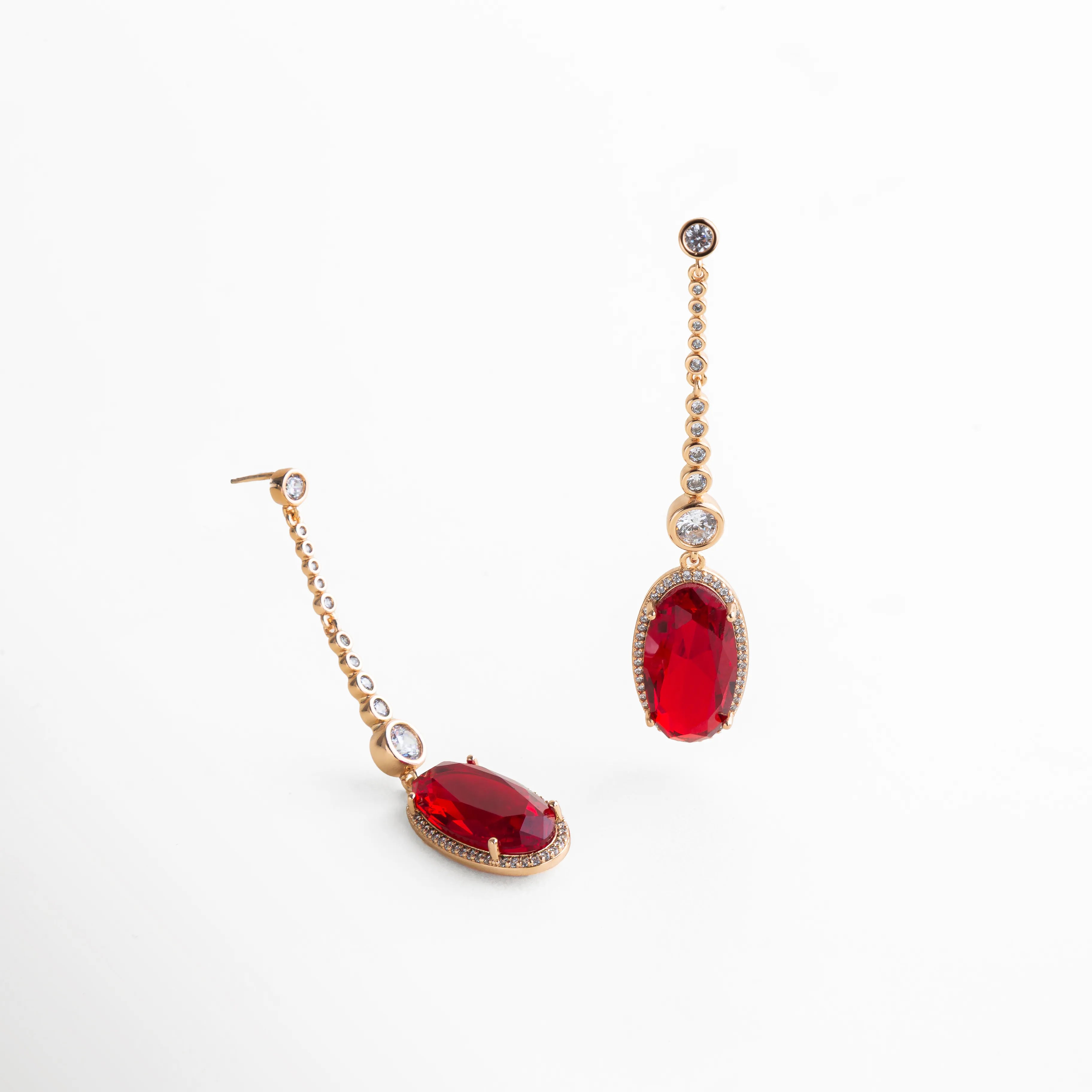 aretes largos dorados con piedra roja ovalada, uno recostado mostrando dimensión y brillo de la piedra, cadena de zirconias, fondo blanco, joyería fina para fiesta