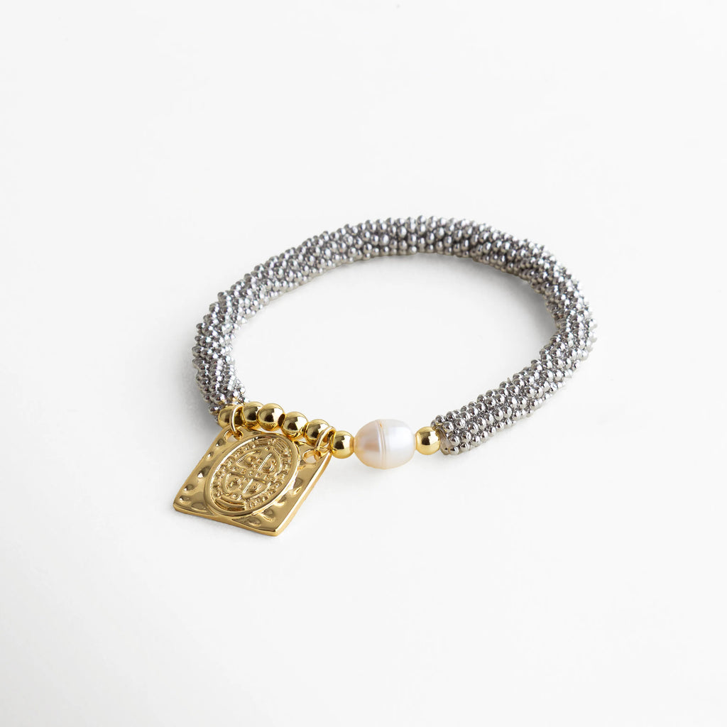 Pulsera metálica plateada con dije dorado religioso San Benito y perla blanca, diseño femenino y sofisticado, perfecta para elevar un look minimalista.