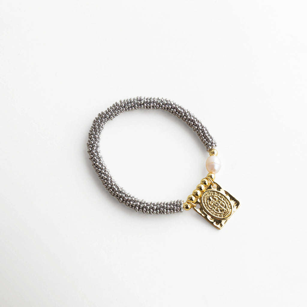 Vista superior de pulsera plateada con dije dorado San Benito y perla, accesorio chic con textura metálica tejida y charm central que da protagonismo.