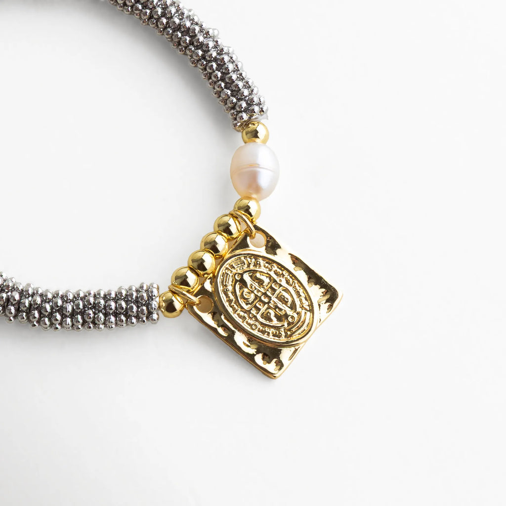 Close up de pulsera plateada con detalle dorado San Benito y perla natural, textura tejida brillante en contraste con un charm espiritual de acabado pulido.