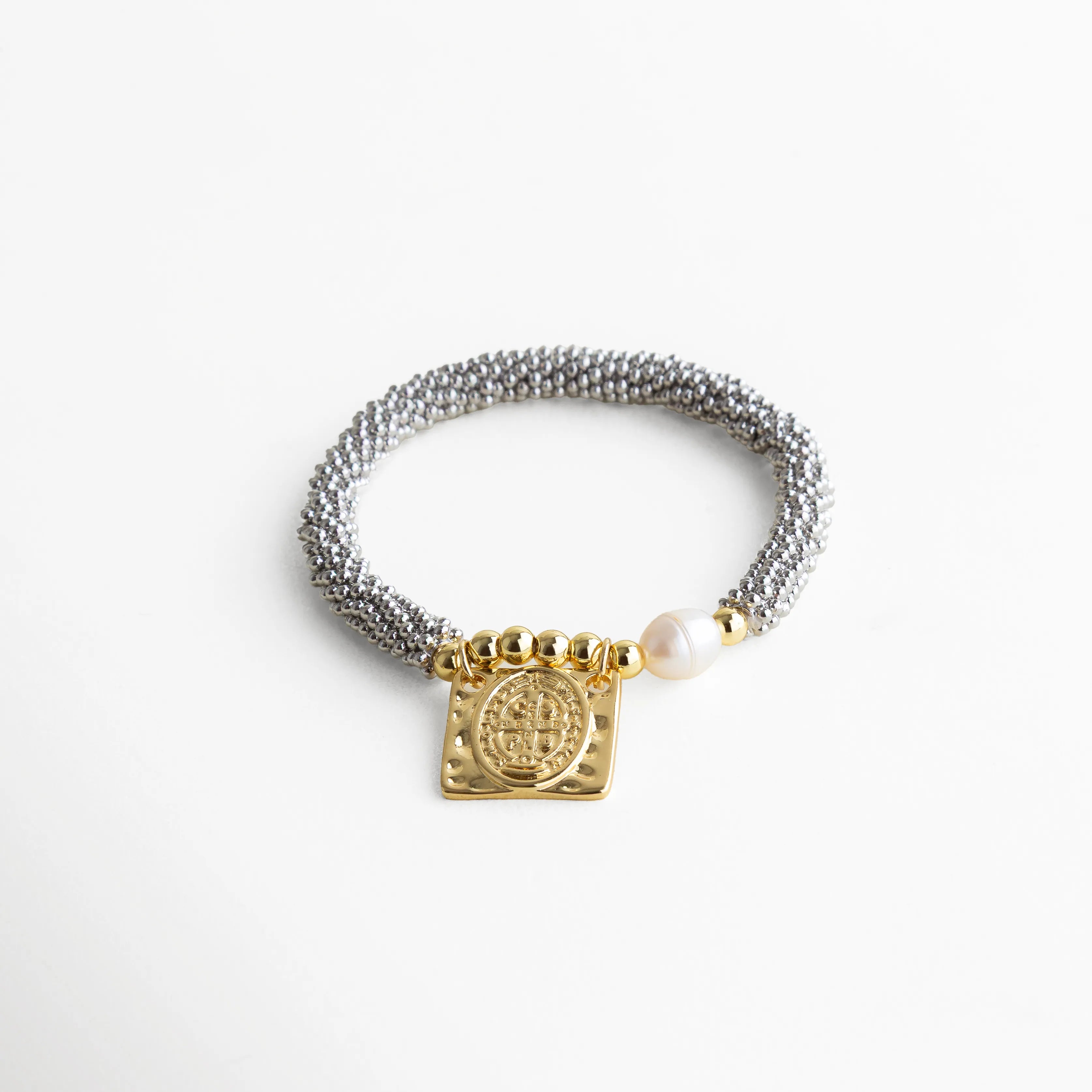 Pulsera plateada tejida con dije dorado San Benito y perla cultivada al frente, estilo elegante y protección espiritual, pieza artesanal que combina tonos metálicos con acentos cálidos.