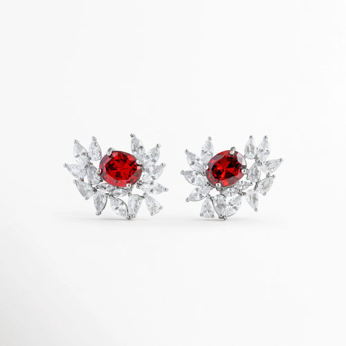 Aretes de topo con rubí sintético ovalado central rodeado de circonias en corte marquesa con diseño de alas o ráfaga brillante, Par de aretes de latón enchapado con gema central roja tipo rubí y marco asimétrico de circonias cristalinas, Vista frontal de aretes elegantes con piedra rubí roja ovalada y halo en forma de abanico con circonias brillantes, Pendientes de topo de 2.5 x 2 cm con piedra central roja intensa y cristales de circonia en disposición radiante.