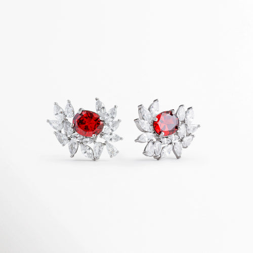 Aretes con centro de rubí ovalado sintético y destellos de circonia en halo asimétrico sobre fondo blanco, Vista de frente de par de pendientes enchapados con piedra roja y cristales transparentes de corte marquesa, Aretes de diseño botánico o de ráfaga con gema ovalada roja y circonias cristalinas brillantes, Par de topos elegantes con rubí sintético central rojo pasión encuadrados en cristales de circonia.