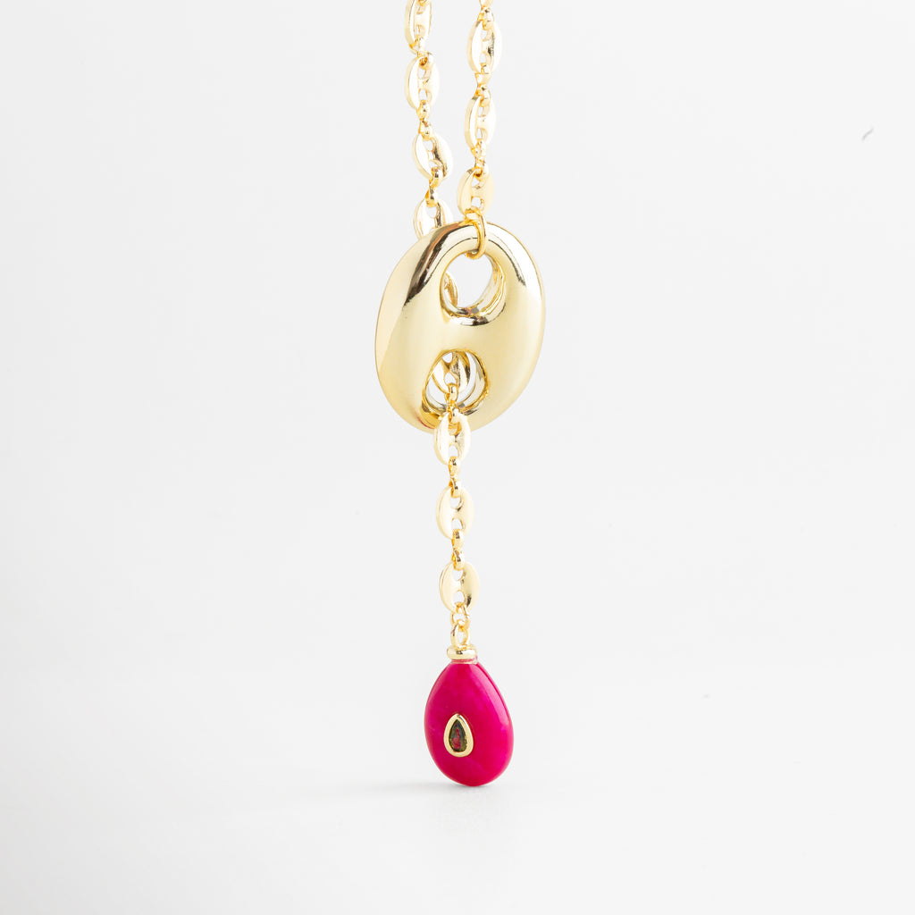 Vista frontal del collar Liriana suspendido mostrando la caída de la gota fucsia, Diseño tipo corbatero: collar dorado con elemento central y piedra rosa colgante, Silueta del collar Liriana resaltando la incrustación de zirconia en la piedra central, Contraste de color: metal dorado y gota fucsia con detalle de zirconia miniatura, Fotografía de producto: collar estilizado con piedra de color y brillo dorado.