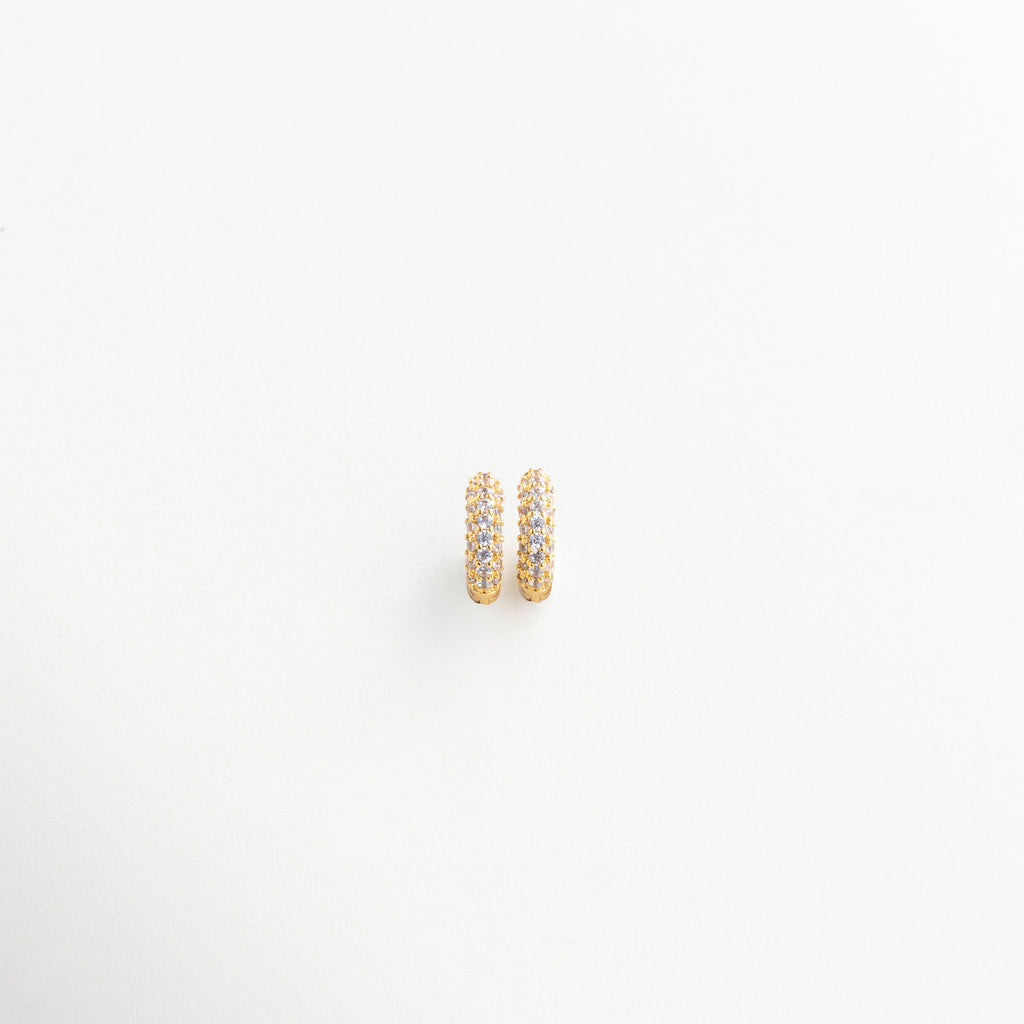 Aros mini vistos en ángulo con detalle de zirconias, Vista lateral de pendientes dorados tipo círculo, Diseño curvo de arracadas pequeñas con brillo sutil, Mini hoops elegantes con textura brillante