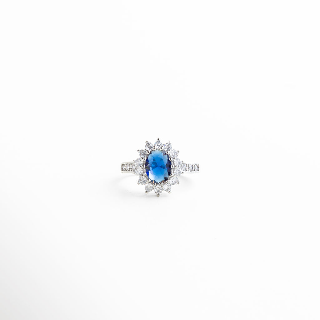 centro ovalado azul con marco de zirconias, engarzado fino con diamantes y piedra central azul, detalles brillantes que rodean la zirconia azul, textura pulida del aro con incrustaciones delicadas, brillo facetado del centro azul profundo