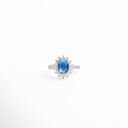 centro ovalado azul con marco de zirconias, engarzado fino con diamantes y piedra central azul, detalles brillantes que rodean la zirconia azul, textura pulida del aro con incrustaciones delicadas, brillo facetado del centro azul profundo