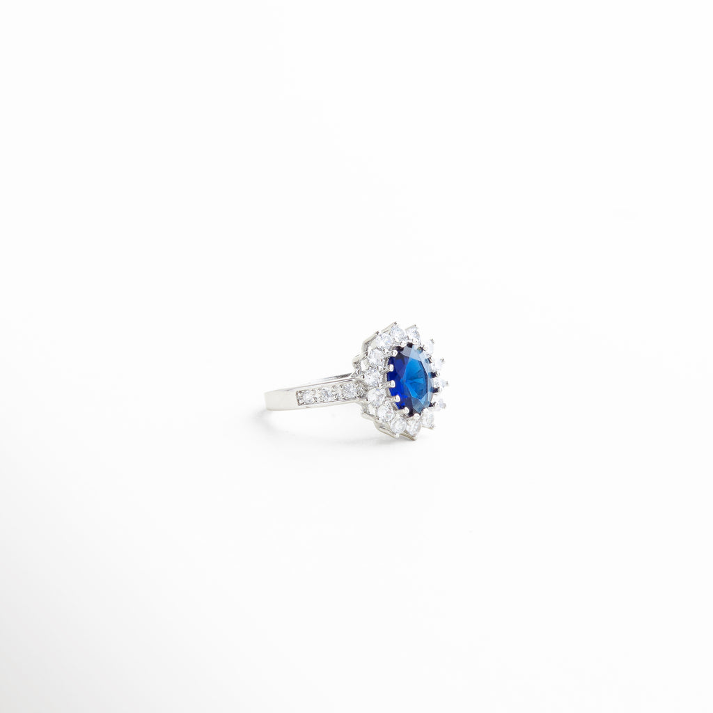 anillo con zirconia azul ovalada al centro, anillo elegante con piedras brillantes y centro azul, diseño clásico con centro color zafiro y detalles brillantes, anillo statement con halo de zirconias y piedra azul, joya con inspiración vintage en tono brillante