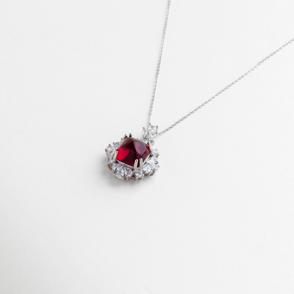 enfoque en la zirconia rubí central, corte preciso que maximiza el brillo, engaste detallado y pulido, contraste entre piedra roja y halo cristalino, diseño que equilibra sutileza y glamour