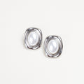 Aretes Oracle Pearl Silver de acero inoxidable con perla central y marco plateado orgánico, Pendientes plateados de estilo vintage con perla sintética blanca en formato ovalado, Vista detallada de aretes tipo "button" plateados con incrustación de perla clásica, Joyería de acero inoxidable: aretes Oracle con acabado plateado y detalle de perla elegante, Aretes de diseño minimalista y elegante con perla blanca y estructura de acero plateado.