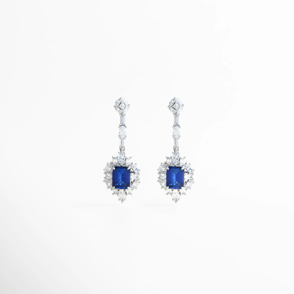 Aretes Cordelia Azules de Plata Ley 925 con piedra central azul zafiro cultivada y halo de cristales, Pendientes largos de plata con gema azul rectangular y detalles de circonias brillantes, Vista frontal de aretes de gala en plata con piedras preciosas de laboratorio color azul real, Joyería fina: aretes Cordelia con diseño de gota y cristales blancos sobre fondo neutro, Aretes elegantes de 4.2 cm con gema azul central, ideales para eventos de noche o bodas.
