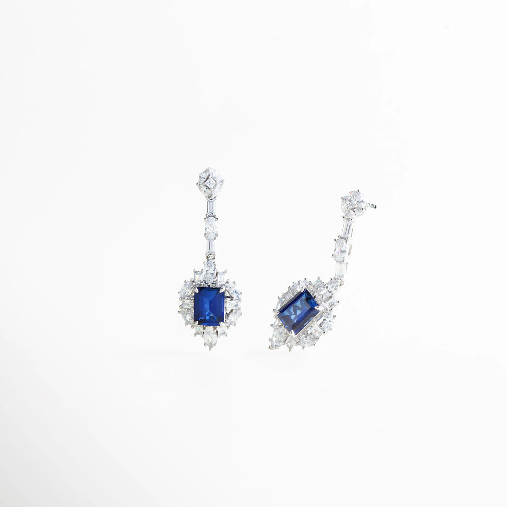 Detalle lateral de los aretes Cordelia resaltando el corte de la piedra azul y el brillo de los cristales, Vista en ángulo de pendientes de plata 925 con gemas azules de laboratorio y acabado pulido, Pendientes con caída elegante y piedras preciosas cultivadas, resaltando el volumen de la pieza, Perspectiva angular de joyería de lujo: aretes con halo de cristales y piedra central azul oscuro, Detalle del engaste de precisión en los aretes Cordelia Azules de plata de ley.