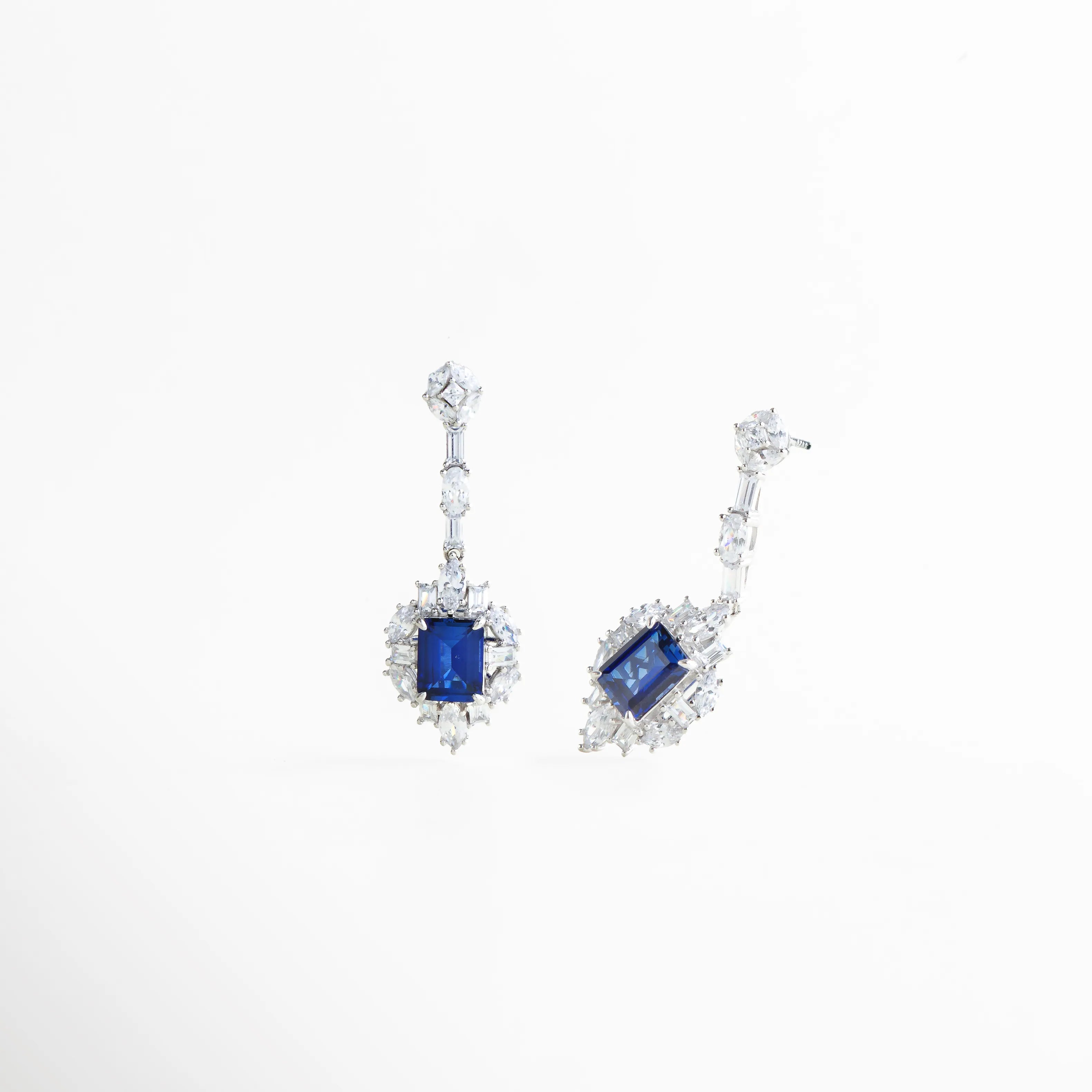 Detalle lateral de los aretes Cordelia resaltando el corte de la piedra azul y el brillo de los cristales, Vista en ángulo de pendientes de plata 925 con gemas azules de laboratorio y acabado pulido, Pendientes con caída elegante y piedras preciosas cultivadas, resaltando el volumen de la pieza, Perspectiva angular de joyería de lujo: aretes con halo de cristales y piedra central azul oscuro, Detalle del engaste de precisión en los aretes Cordelia Azules de plata de ley.