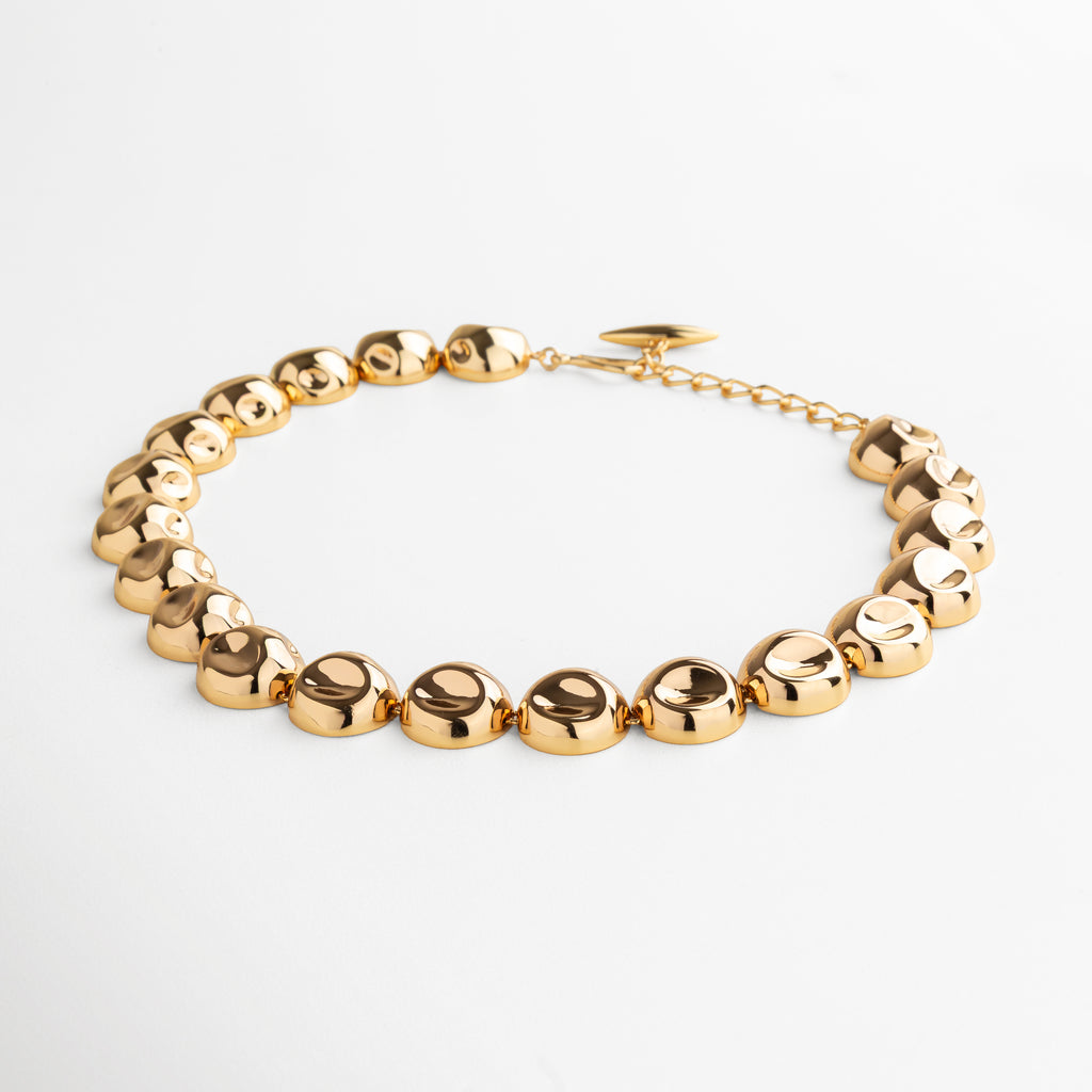 Collar Golden Bell dorado visto de perfil, mostrando la forma y grosor de los dijes unidos. Vista lateral del collar con dijes circulares, resaltando la continuidad y acabado pulido en oro 24K. Perfil del collar con diseño de dijes unidos, ideal para un look moderno y sofisticado. Detalle lateral del Collar Golden Bell, captando la textura y el brillo uniforme de los dijes. Collar con dijes circulares en acabado dorado visto desde un ángulo lateral, mostrando su estructura y estilo.