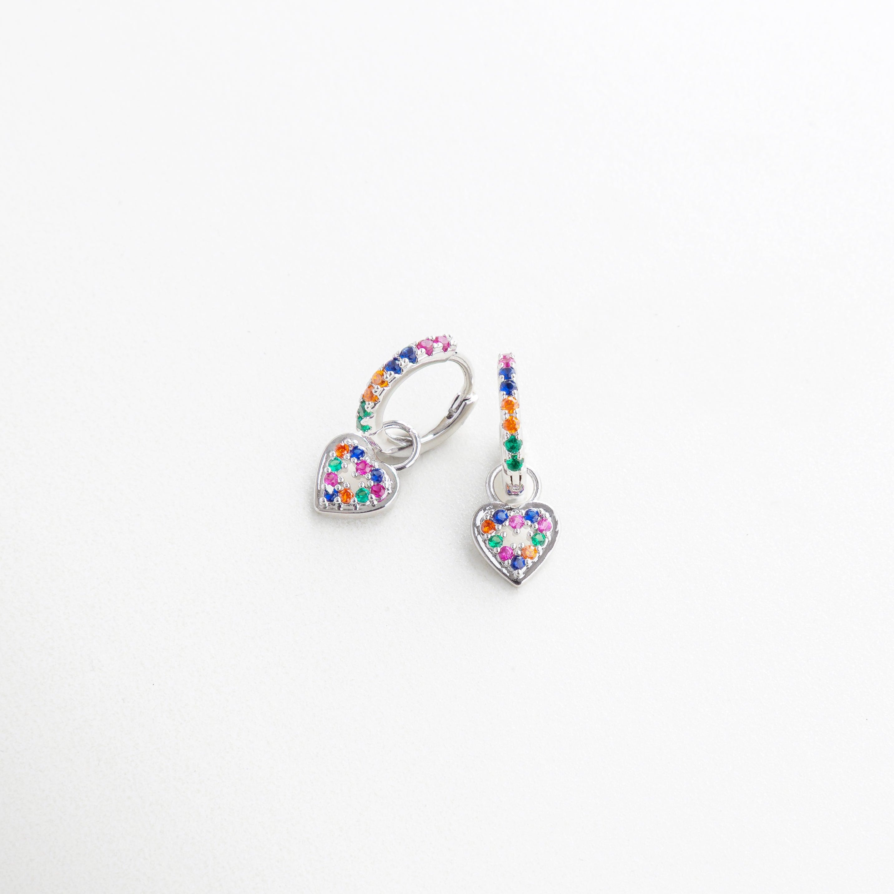 Aretes plateados con corazón colgante vistos de perfil, Diseño curvado con charm colorido en perspectiva, Arracadas en ángulo con piedras de colores, Pendientes plateados con detalle romántico