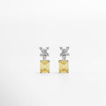 Aretes plateados con piedra amarilla cuadrada al centro, vistos de frente sobre fondo neutro. Vista frontal de aretes elegantes en tono plateado, con piedra amarilla brillante en corte cuadrado. Aretes rectangulares plateados con centro amarillo, estilo refinado y sofisticado. Primer plano frontal de par de aretes plateados con piedra amarilla en diseño clásico y elegante.Aretes plateados con incrustación cuadrada amarilla, vistos de frente con acabado brillante.