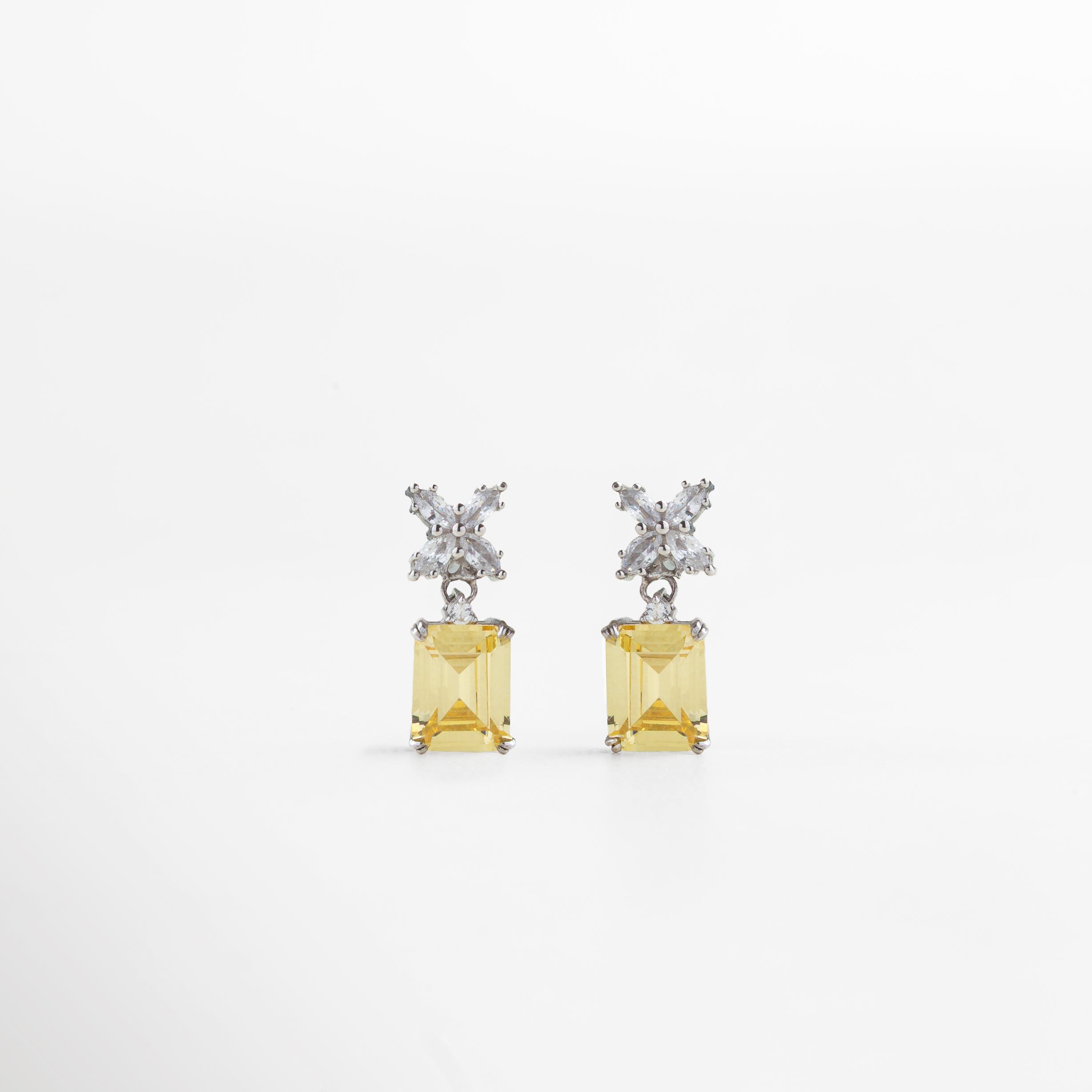 Aretes plateados con piedra amarilla cuadrada al centro, vistos de frente sobre fondo neutro. Vista frontal de aretes elegantes en tono plateado, con piedra amarilla brillante en corte cuadrado. Aretes rectangulares plateados con centro amarillo, estilo refinado y sofisticado. Primer plano frontal de par de aretes plateados con piedra amarilla en diseño clásico y elegante.Aretes plateados con incrustación cuadrada amarilla, vistos de frente con acabado brillante.