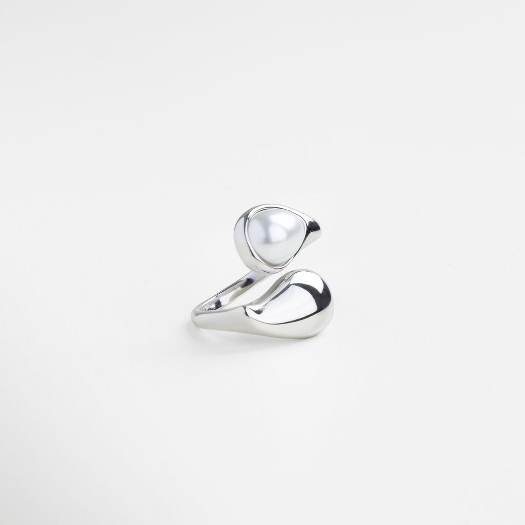 Anillo con perla en forma ondulada desde ángulo lateral, Perfil escultórico del anillo plateado con perla, Diseño envolvente en plateado con perla central, Curvas modernas en plata con perla elevada