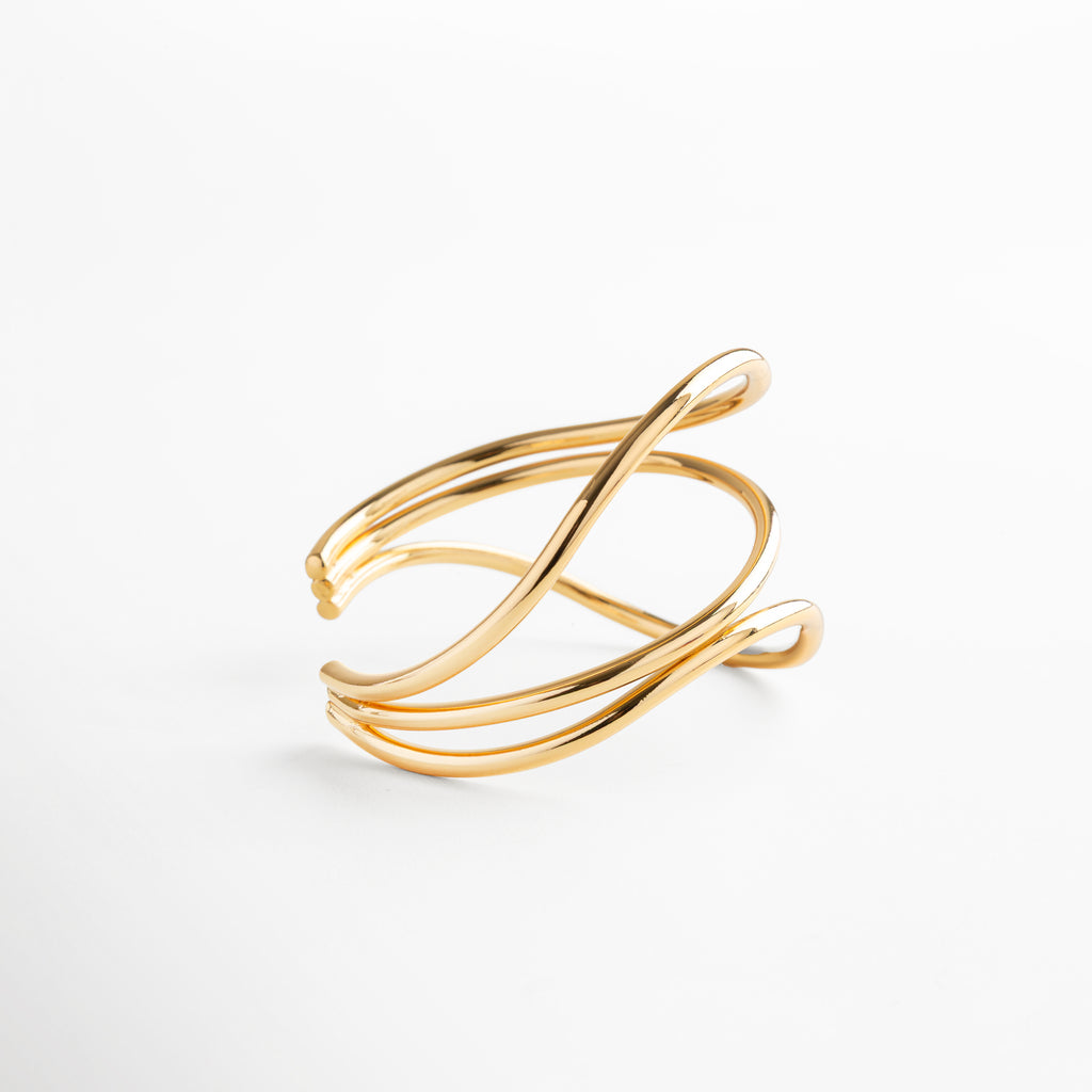 Pulsera dorada con forma escultórica y líneas curvas, acabado espejo en oro 24K, accesorio atemporal que equilibra movimiento y elegancia.